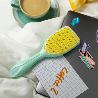 Grande brosse démêlante Agamy vert pastel et picots jaunes, posée sur un lit blanc avec ordinateur , une tasse de café.