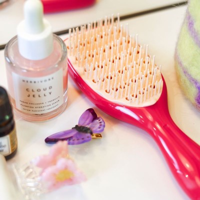 Petite brosse démêlante Agamy framboise avec picots rose clair, posée parmi des soins et accessoires