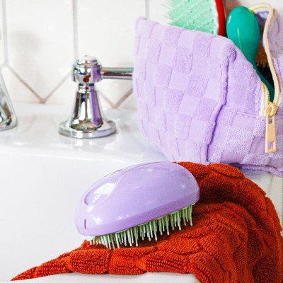 Brosse compacte AGAMY violette posée sur une serviette rouge dans une salle de bain colorée, trousse assortie en arrière-plan